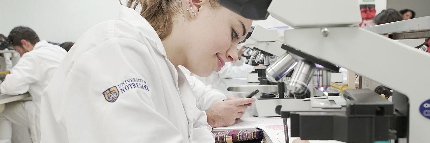 Students wearing white lab coats with University of Notre Dame seal using microscopes.