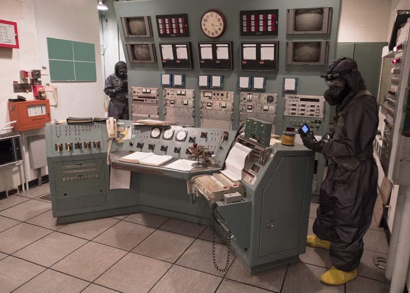 Control room filled with dials and gauges with two soldiers wearing full MOPP protective gear, gloves, boots, gloves and using Geiger counters. Biography of Dr. Anthony J. Carbone.